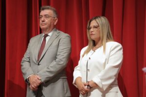 Two individuals stand side by side in front of a red curtain, both looking serious. One wears a light gray suit, the other a white blazer.