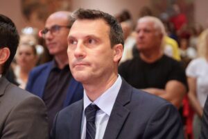 A man in a suit looks thoughtfully at an event, surrounded by an audience in a blurred background.