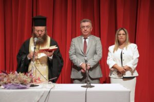 A man in religious attire reads from a book at a podium, flanked by two individuals in formal wear, against a red curtain backdrop.