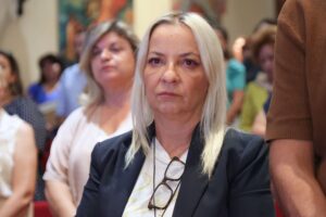A woman with long, straight blonde hair sits in a crowd, looking serious. Others are blurred in the background.