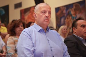 A man in a blue shirt sits among an audience, looking serious, with colorful artwork in the background.