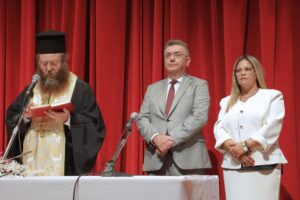 A man in religious attire reads from a book, flanked by two individuals in formal clothing, against a red curtain backdrop.