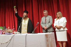 A man in religious attire holds a green object, flanked by two individuals, in front of a red curtain at a formal event.