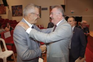 Two men in suits are smiling and shaking hands in a formal setting, with an audience in the background.