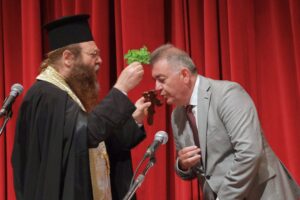 A clergy member in traditional attire blesses a man during a ceremony, with a red curtain in the background.