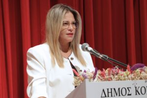 A woman with long hair speaks at a podium adorned with flowers, set against a red curtain backdrop.