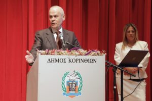 A man speaks at a podium with a floral arrangement, while a woman stands beside him, in front of a red curtain.