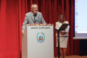A man in a gray suit speaks at a podium with a woman in a white outfit standing beside him, in front of a red curtain.