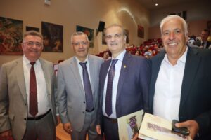 Four men in formal attire pose together in a hall, smiling and holding books. The background features colorful artwork.