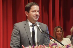 A man in a suit speaks at a podium with a microphone, set against a red curtain backdrop. Flowers decorate the podium.