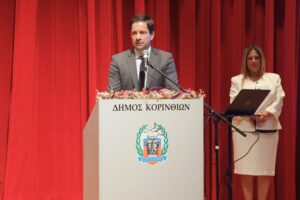 A man in a suit speaks at a podium with a seal, while a woman in a white outfit stands beside him. Red curtains in the background.