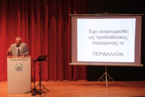 A speaker presents at a podium with a screen displaying text in Greek about the environment. Red curtains in the background.