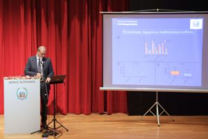 A speaker stands at a podium with a presentation screen behind, displaying graphs and data, in front of red curtains.