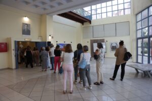 A group of people stands in line inside a spacious, well-lit lobby with large windows and a tiled floor.