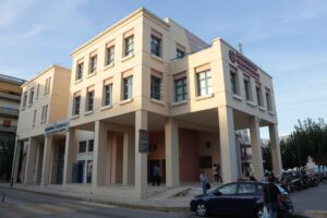 A modern building with a light-colored facade, featuring multiple windows and a raised entrance. Vehicles are parked nearby.