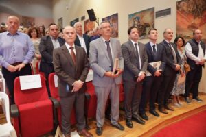 A group of people standing in a formal setting, some holding books, with a backdrop of artwork and red seating.