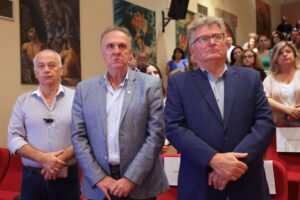 Three men stand in a formal setting, looking serious, with a crowd and artwork in the background.