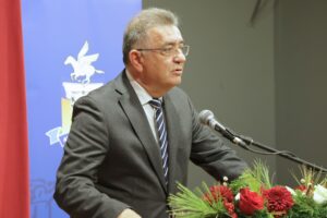 A man in a suit speaks at a podium with a microphone, surrounded by flowers, against a blue backdrop.