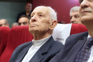 An elderly man with white hair sits in a red chair, looking thoughtfully ahead, surrounded by other attendees at an event.