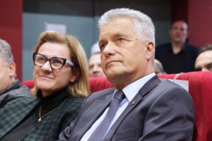 A man in a suit and a woman with glasses sit together, both looking thoughtfully at an event.