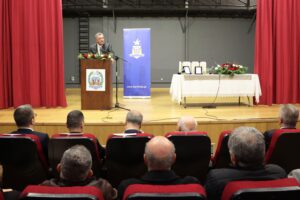 A speaker addresses an audience in a theater setting, with a podium and a table displaying framed items and flowers.