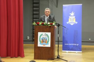 A speaker at a podium with flowers, addressing an audience in a hall, with a blue banner in the background.