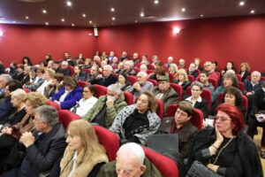A large audience seated in a theater, with individuals of various ages and styles, against a red wall backdrop.