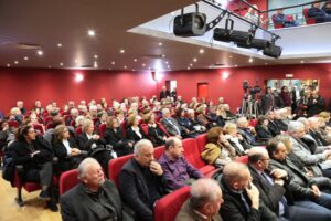 A large audience seated in a theater, with many people in formal attire, facing a stage area. The room has red walls and bright lighting.