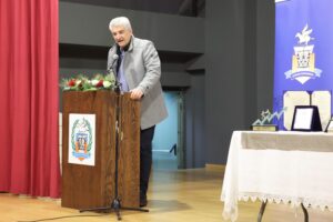 A man speaks at a podium decorated with flowers, with awards displayed on a table nearby. Red curtains and a blue backdrop are visible.