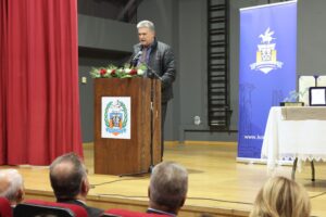 A speaker addresses an audience from a podium adorned with flowers, with a banner in the background. Red curtains frame the scene.