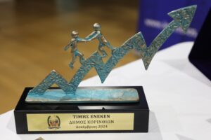 A decorative trophy featuring two figures climbing a stylized upward arrow, with a plaque inscribed in Greek.