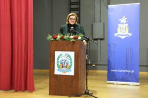 A woman stands at a podium with flowers, speaking at an event. A blue banner with a logo is in the background.