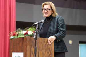 A woman with glasses stands at a podium decorated with flowers, speaking into a microphone. Red curtains are in the background.