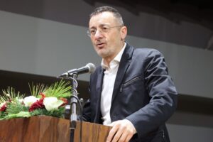 A man in a black suit speaks at a podium adorned with flowers, delivering a speech in a well-lit venue.