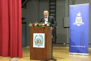 A man speaks at a podium decorated with flowers, in front of a backdrop featuring a logo and red curtains.