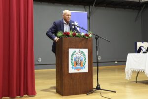 A man stands at a podium with a floral arrangement, speaking at an event. A table with a white cloth is visible nearby.