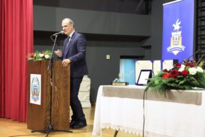 A man in a suit speaks at a podium with a microphone, surrounded by awards and floral arrangements on a table.
