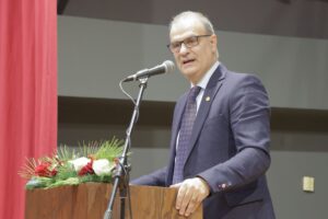 A man in a suit speaks at a podium decorated with flowers, with a microphone in front of him. Red curtains are in the background.
