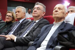 A group of four people seated in a theater, attentively watching an event. They appear engaged and serious.