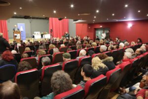 A large audience seated in a theater, facing a speaker at a podium, with red curtains in the background.