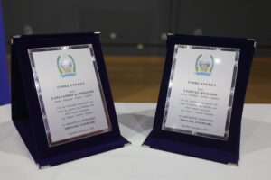 Two framed awards displayed on a table, featuring names and logos, set against a blurred background.