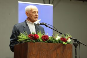 An elderly man speaks at a podium adorned with flowers, with a blue backdrop behind him.