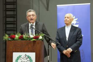 Two men are speaking at a podium adorned with flowers. One is gesturing while the other stands beside him, listening.