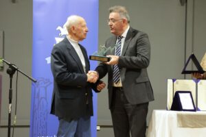 Two men are shaking hands on stage, one receiving an award. A blue backdrop and a table with certificates are visible.