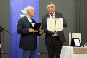 Two men stand together at an event, one holding a certificate and the other a medal, with a blue backdrop and awards on a table.