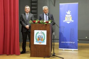 A man speaks at a podium with a floral arrangement, while another man stands beside him. A banner is displayed in the background.