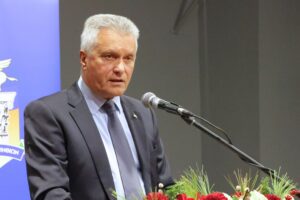 A man in a suit speaks at a podium with a microphone, set against a backdrop featuring a logo. Flowers are arranged in front.