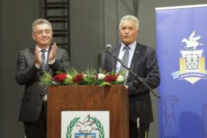 Two men stand at a podium with flowers, one speaking and the other applauding. A banner is visible in the background.