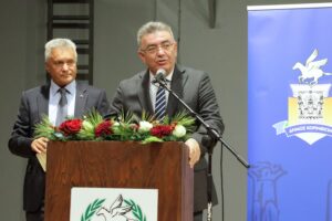 Two men are speaking at a podium decorated with flowers, with a blue banner in the background.