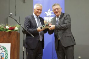 Two men are smiling and exchanging a trophy at a formal event, with a podium and a blue backdrop in the background.